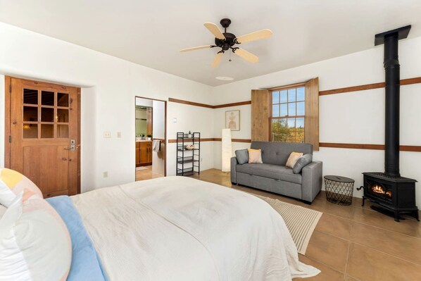 A bedroom with a bed, a grey sofa, a wooden door, a window, a ceiling fan, and a wood-burning stove.