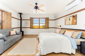 A bedroom with a bed, a sofa, a window with a view, a ceiling fan, and an air conditioning unit.