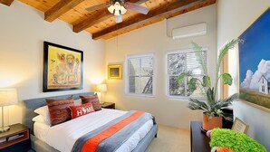 Un dormitorio con cama, ventilador de techo, ventana con persianas, planta en maceta e imagen enmarcada en la pared.