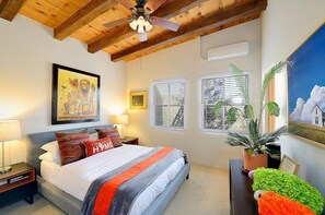 Ein Schlafzimmer mit Bett, Deckenventilator, Fenster mit Jalousien, Topfpflanze und einem gerahmten Bild an der Wand.