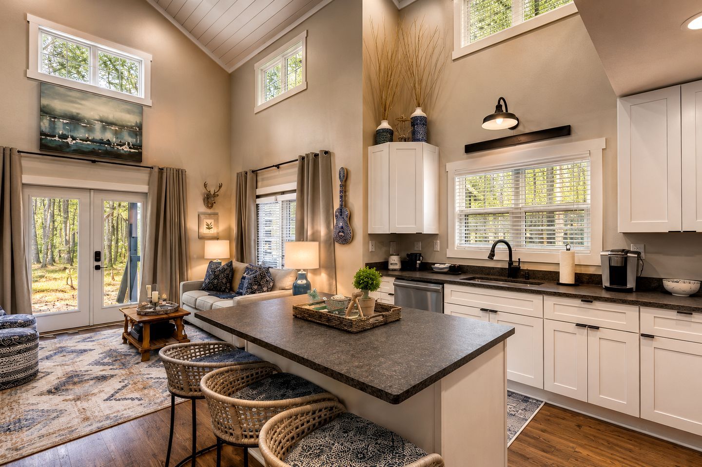 A modern kitchen with a central island, bar stools, a dining area with a sofa, and a view of the outdoors through large windows.