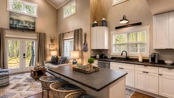 A modern kitchen with a central island, bar stools, a dining area with a sofa, and a view of the outdoors through large windows.