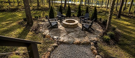 Un foyer en pierre, entouré de fauteuils Adirondack, dans un cadre forestier.