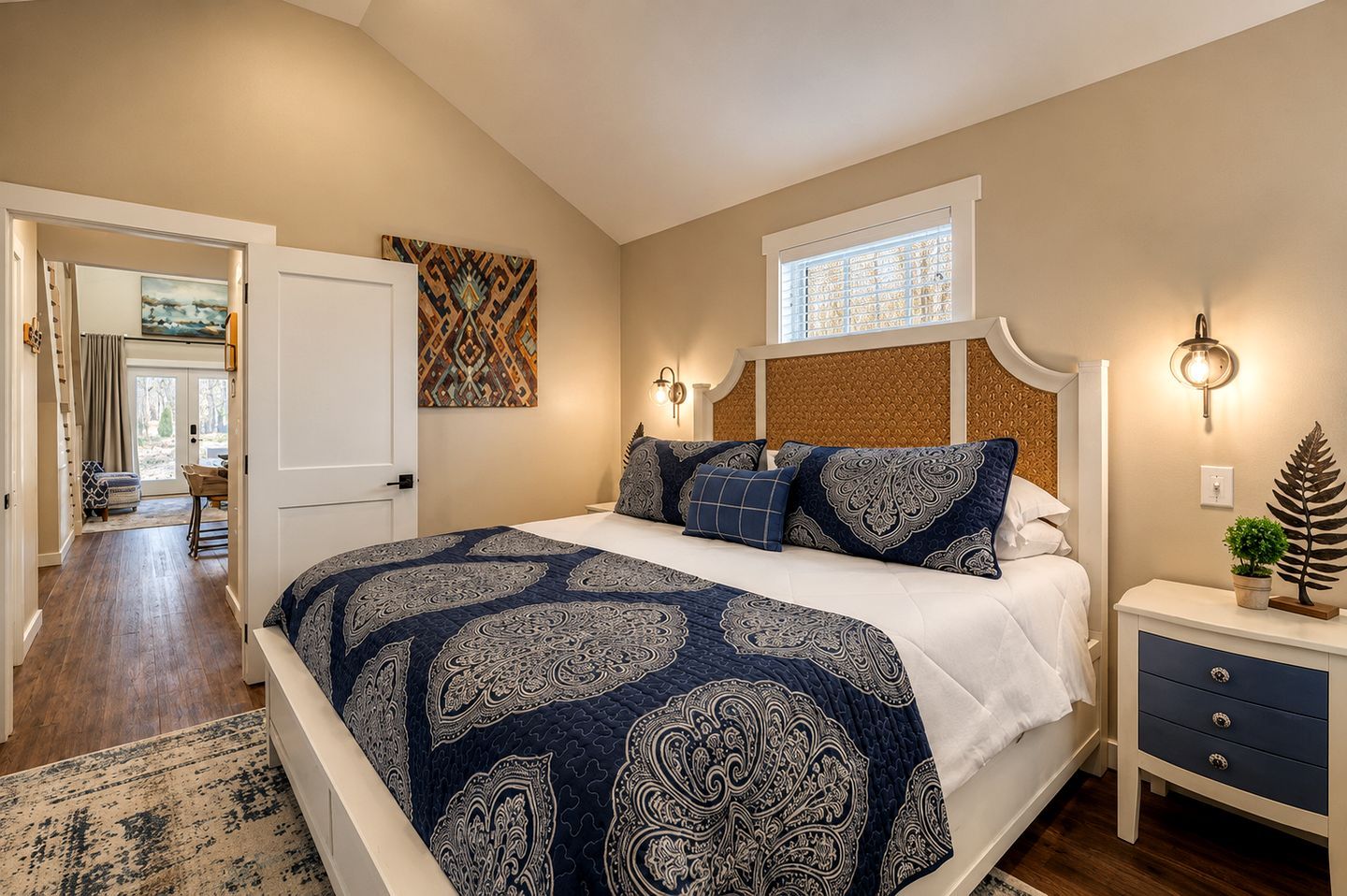A bedroom with a large bed, a nightstand, a window with blinds, and a door leading to another room.