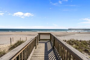 Ein Holzsteg führt zu einem Strand, in der Ferne ist ein Steg zu sehen.