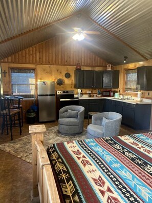 Un chalet rustique comprenant une cuisine, deux fauteuils, une table à manger et un grand lit recouvert d’une couverture à motifs.