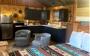 Une cuisine compacte équipée d’un réfrigérateur, d’une cuisinière, d’un four à micro-ondes et de meubles de rangement sombres. On y trouve deux fauteuils et un lit recouvert d’une couverture à motifs.