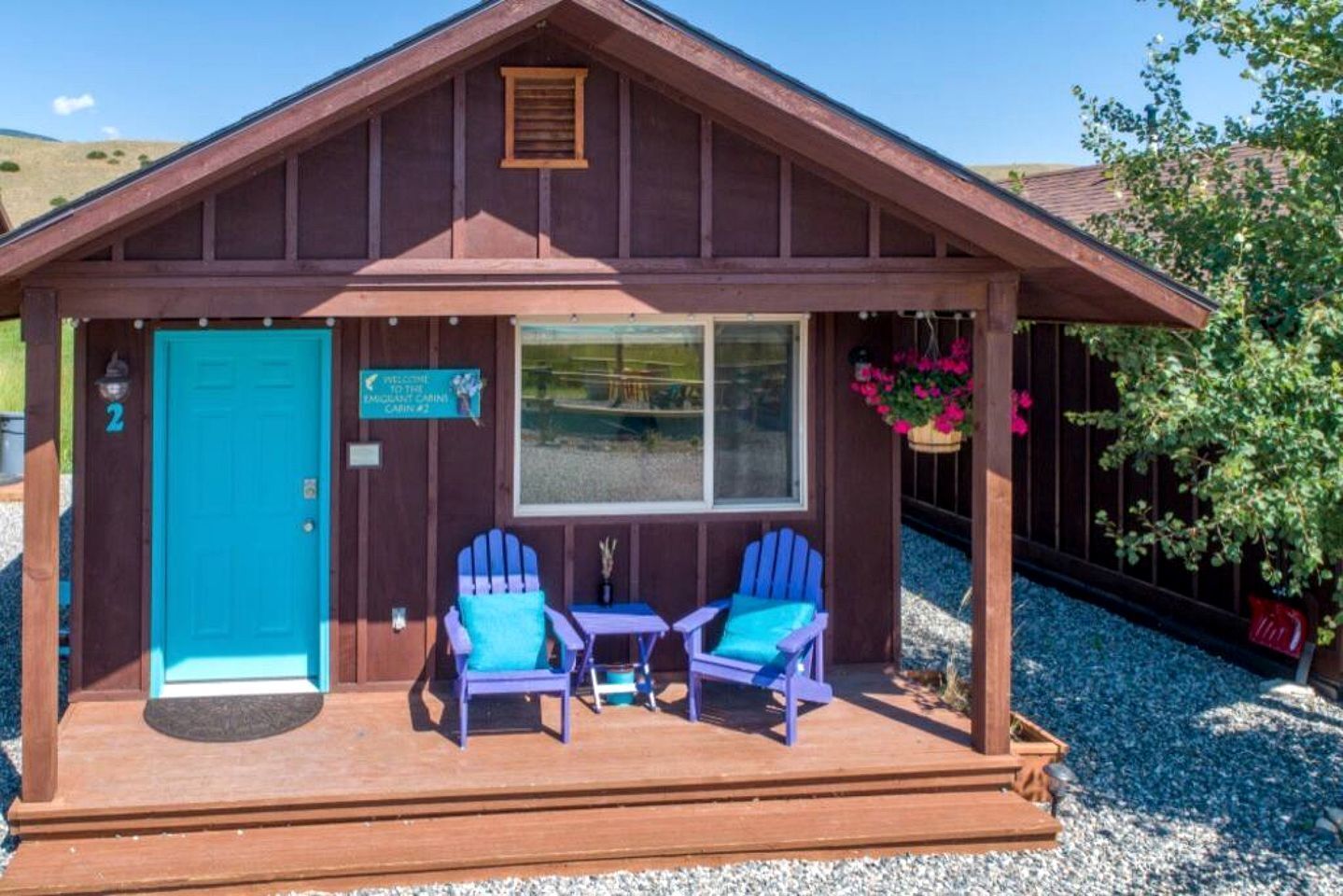 Una cabaña con un porche, dos sillas Adirondack, una mesita, una puerta azul y un cartel de bienvenida.