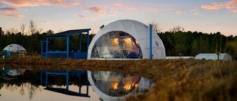 Una estructura abovedada con techo de cristal, rodeada por un pabellón azul y reflejada en el agua.