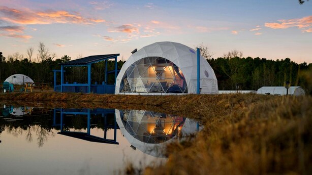 Una struttura a forma di cupola con soffitto in vetro, circondata da un padiglione blu, che si riflette in uno specchio d'acqua.