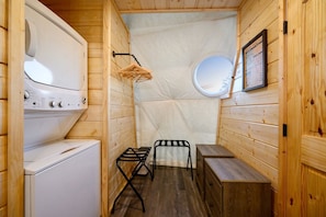 A compact wooden cabin with a washing machine, a drying rack, a small table, and a framed informational poster.