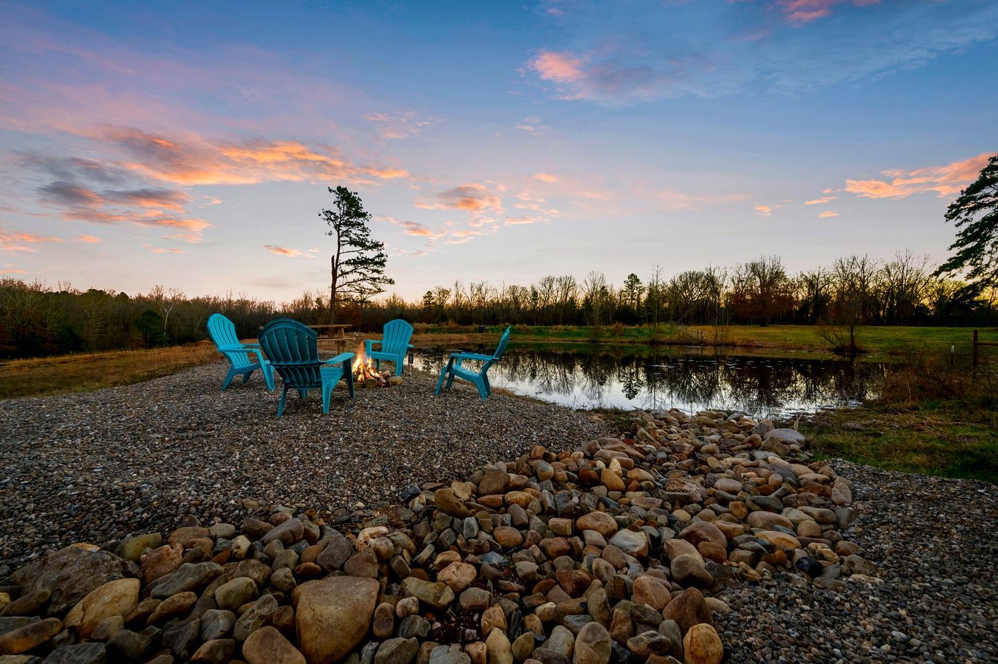A gravel area with three blue Adirondack chairs, a small fire, and a pond.