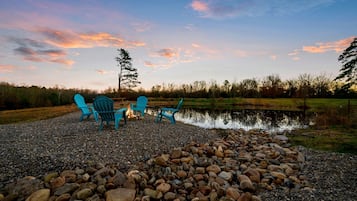 A gravel area with three blue Adirondack chairs, a small fire, and a pond.