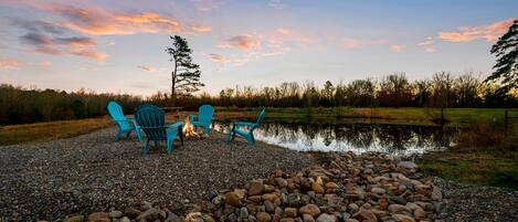 A gravel area with three blue Adirondack chairs, a small fire, and a pond.