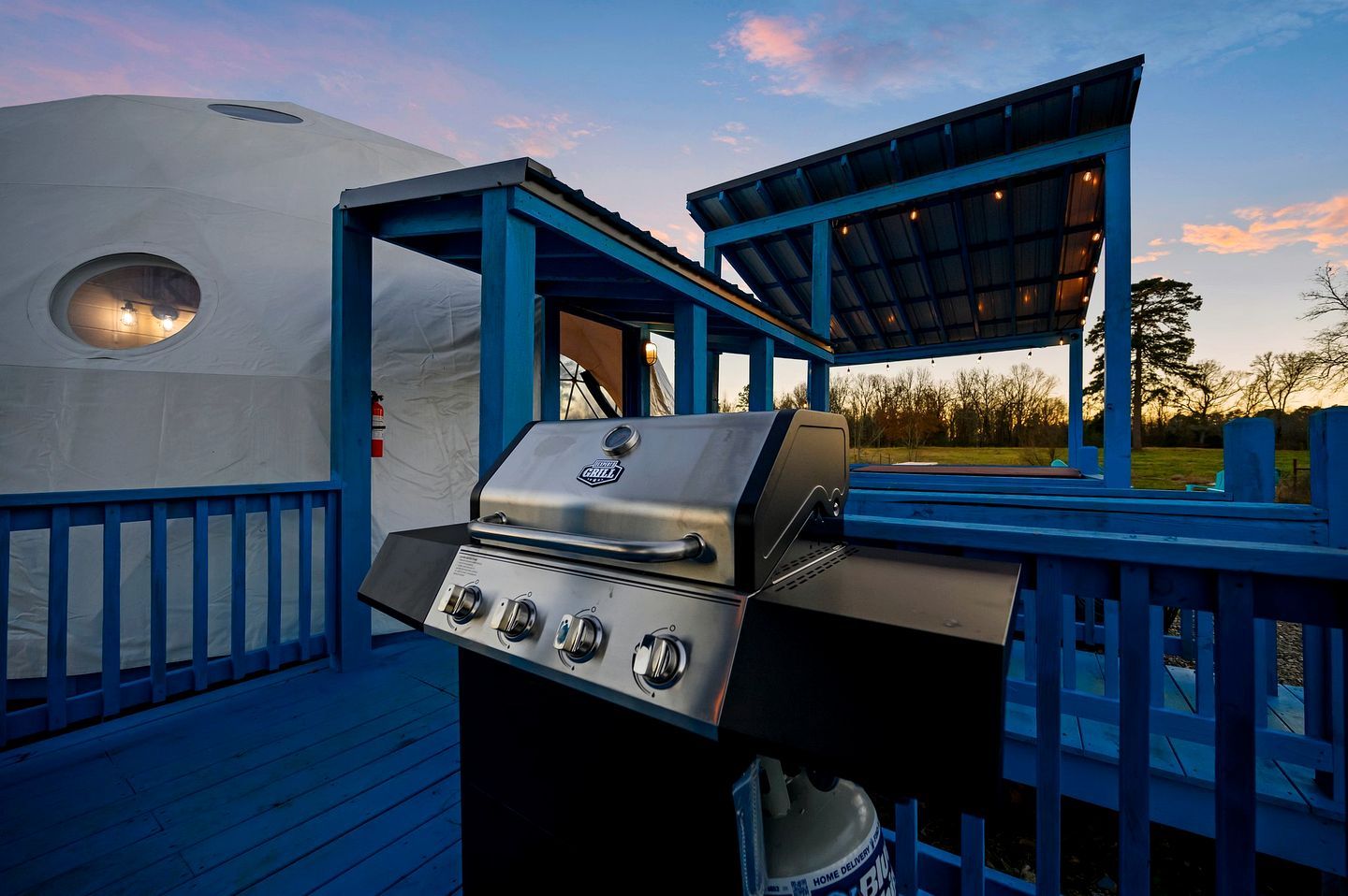 Uma área de churrasco coberta ao ar livre, com estrutura azul, uma tenda e um deck de madeira.