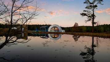Um edifício em forma de cúpula à beira de um lago calmo, com reflexos, cercado por árvores e um céu claro.