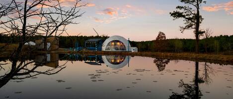 A dome-shaped building by a calm lake with reflections, surrounded by trees and a clear sky.