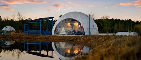 Una estructura abovedada con techo de cristal, rodeada por un pabellón azul y reflejada en el agua.