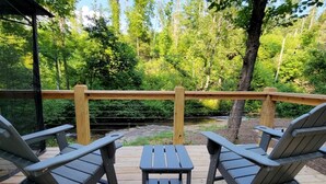 A deck with Adirondack chairs and a small table, overlooking a forested area with a wooden railing. - Geodome on Creek, Great Getaway, WIFI & Hot Shower near Boone, NC (Purlear)