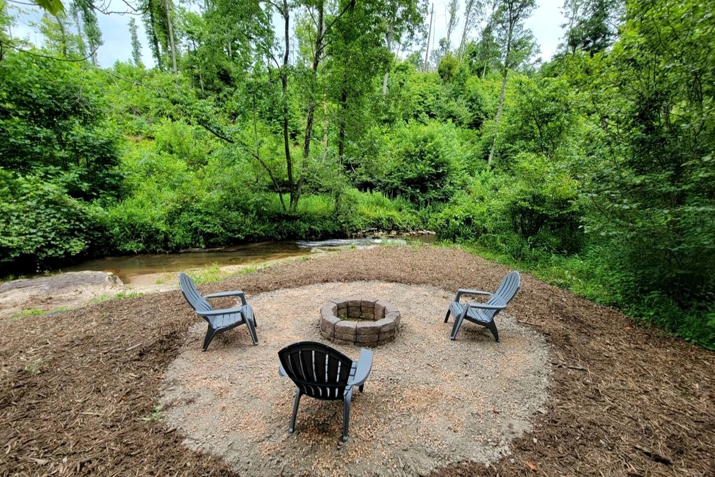 Ein kreisförmiger Sitzbereich mit drei Adirondack-Stühlen um eine Feuerstelle, umgeben von Wald und einem Bach.