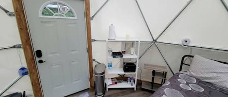 Una habitación con carpa, una cama, un estante blanco con objetos, una puerta y una pequeña mesa.