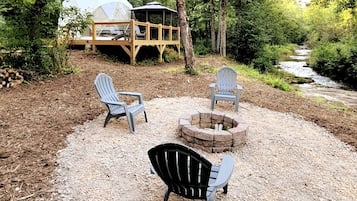 A campsite with a fire pit, two Adirondack chairs, and a covered area with a dome structure.