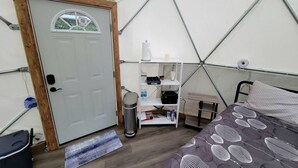 A tented room with a bed, a white shelf with items, a door, and a small table. - Geodome on Creek, Great Getaway, WIFI & Hot Shower near Boone, NC (Purlear)
