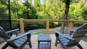 Terraza con sillas tipo Adirondack y una pequeña mesa, con vista a una zona boscosa y barandilla de madera.