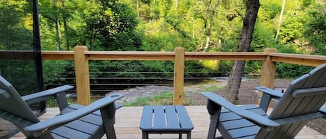 Una terrassa amb cadires Adirondack i una taula petita, amb vista a una zona boscosa i una barana de fusta.