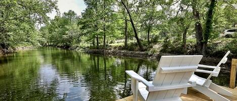 Un muelle de madera con una silla Adirondack blanca, rodeado de árboles y un río tranquilo.
