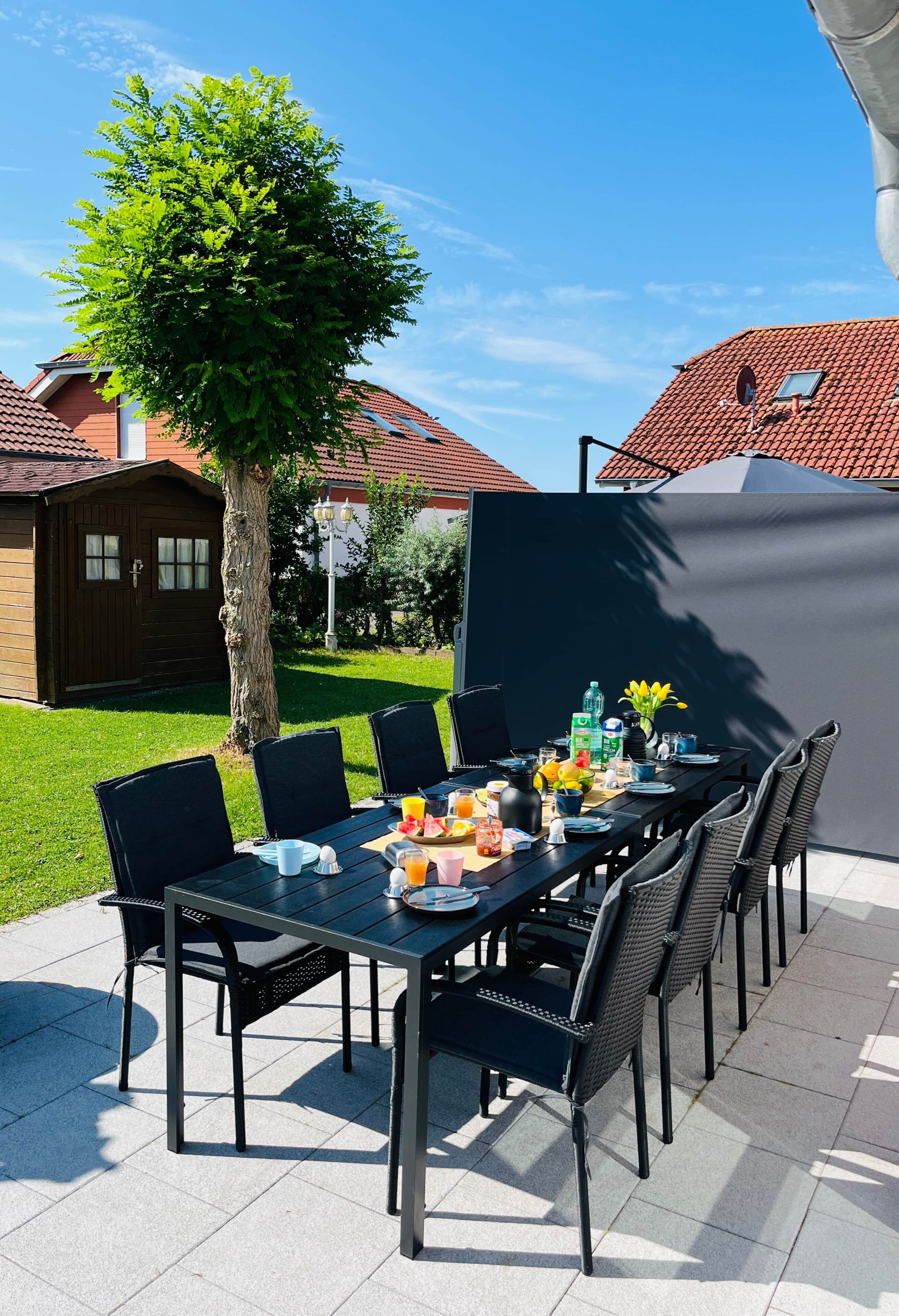 Ein Essbereich im Freien mit einem für das Frühstück gedeckten Tisch, umgeben von Stühlen, einem Baum und einem Gebäude mit roten Ziegeldach.