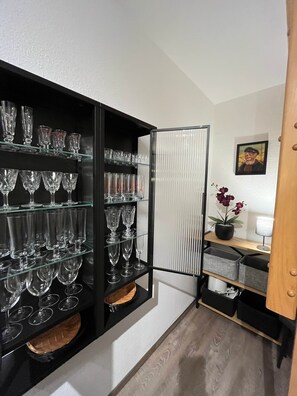 Une pièce de taille modeste, avec une étagère noire remplie de verres à vin, une plante en pot et un tableau encadré accroché au mur.