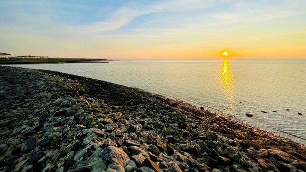 A coastal scene with a rocky shoreline, calm water, and a setting sun. - Lüttje Deichvilla Backbord (Wurster Nordseeküste)