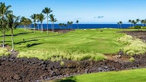 A golf course with a view of the ocean, surrounded by palm trees and volcanic rock formations. - Amazing Ocean, Golf & Sunset Views Great Location Private Bbq! - Vrkcv (Waikoloa)