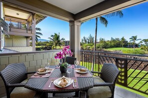 A covered outdoor dining area with wicker furniture, a table set for two, and a view of a well-maintained garden and distant buildings. - Amazing Ocean, Golf & Sunset Views Great Location Private Bbq! - Vrkcv (Waikoloa)