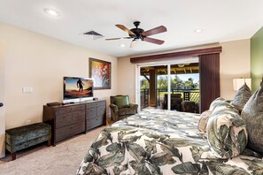A bedroom with a large bed, a ceiling fan, a television, a bench, and a sliding glass door leading to an outdoor area. - Amazing Ocean, Golf & Sunset Views Great Location Private Bbq! - Vrkcv (Waikoloa)
