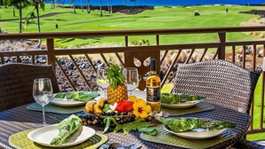 A dining table set with plates, glasses, a bottle, and tropical fruits on a balcony overlooking a golf course and ocean. - Amazing Ocean, Golf & Sunset Views Great Location Private Bbq! - Vrkcv (Waikoloa)