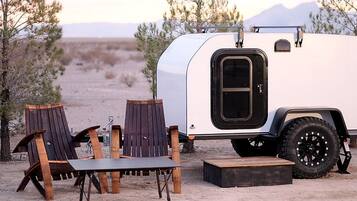 A white RV with a black door and large tires parked in a desert with two wooden Adirondack chairs and a small table.