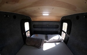 Interior compacto de una autocaravana con una cama individual, una mesita y una ventana.