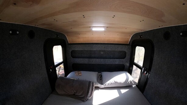 A compact RV interior with a single bed, a small table, and a window.