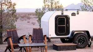 A white RV with a black door and large tires parked in a desert with two wooden Adirondack chairs and a small table.