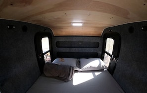 A compact RV interior with a single bed, a small table, and a window.