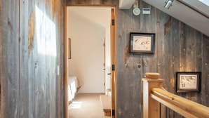 Un escalier en bois avec une rampe, un tableau encadré accroché au mur et une porte donnant sur une pièce où se trouvent un lit et un banc.