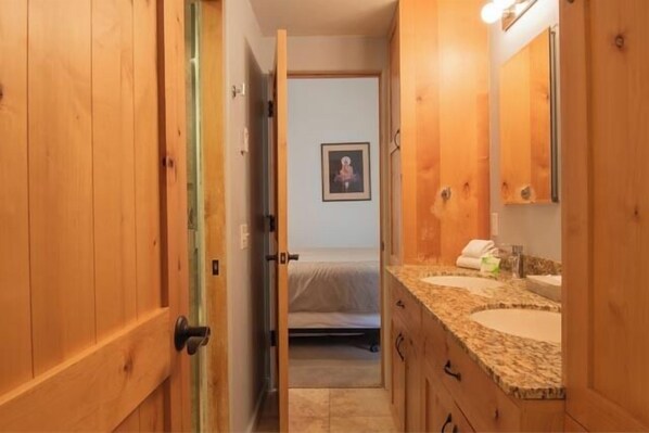Un pasillo estrecho conduce a un dormitorio con una cama y un cuadro enmarcado en la pared. La habitación tiene puertas de madera, una encimera de granito con un lavabo y un espejo.