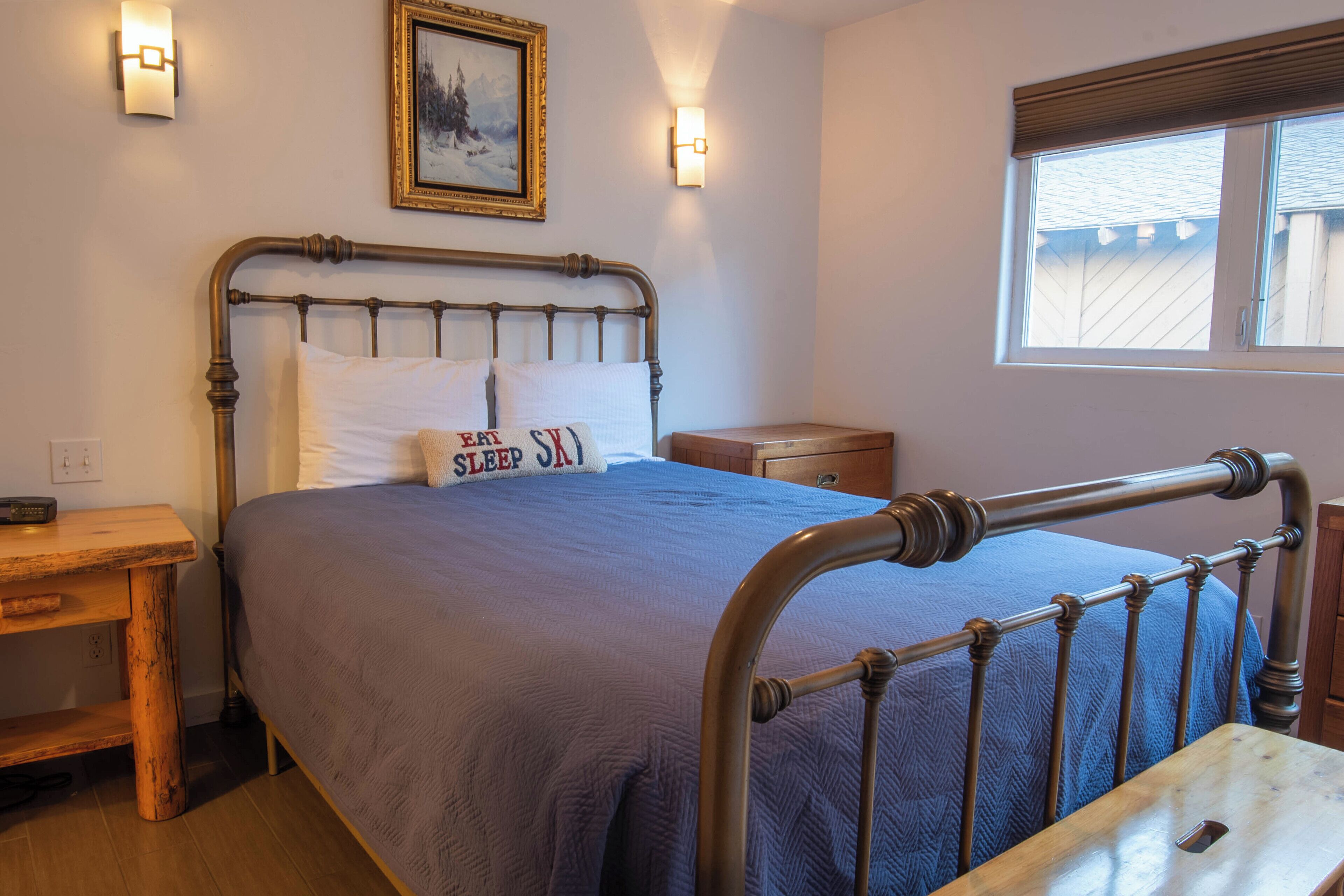 A bedroom with a metal bed frame, a wooden nightstand, a bedside table, and a window with blinds.