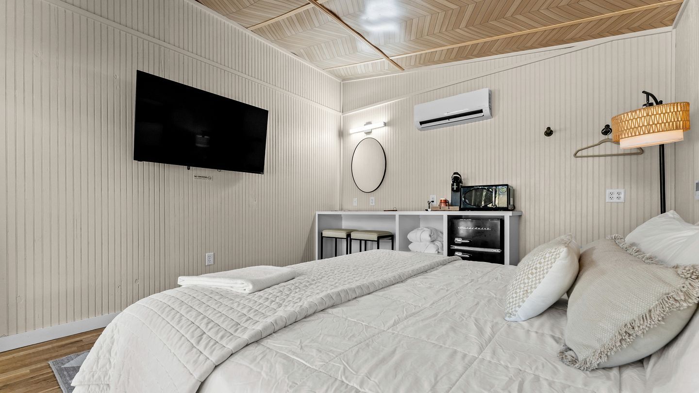 A hotel room with a bed, a television, an air conditioning unit, a desk with a computer, and a lamp.