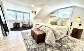 A bedroom with a large bed, a small table, a chair, and a window with blinds.