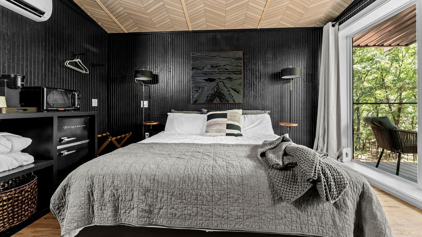 A bedroom with a bed, a side table, a lamp, a painting, and a view of trees through a window.
