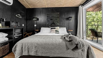 A bedroom with a bed, a side table, a lamp, a painting, and a view of trees through a window.
