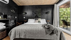 A bedroom with a bed, a side table, a lamp, a painting, and a view of trees through a window.